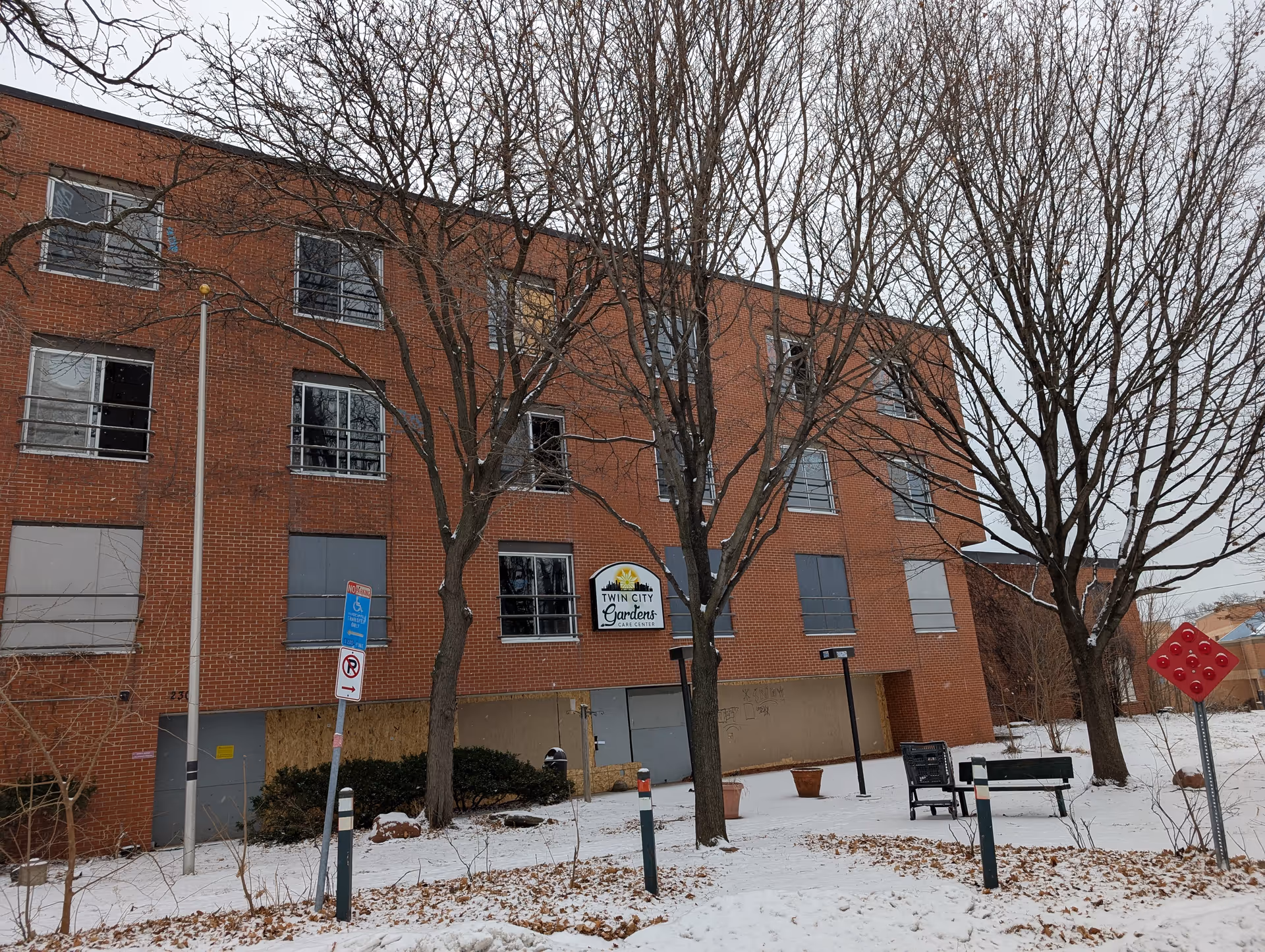 Exterior view of a three-story brick building in winter with leafless trees and snow on the ground. Several windows are boarded up or broken. A sign on the building reads 'Twin City Gardens Care Center'. There are parking signs and a bench in front of the building.