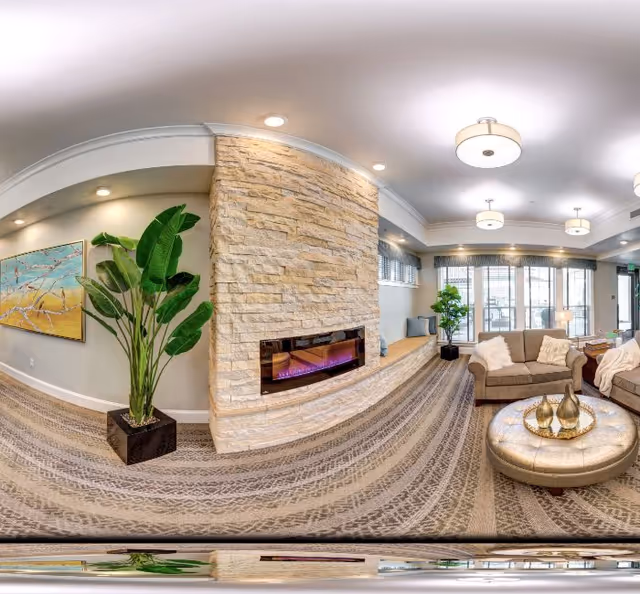 A spacious and well-lit living room area featuring a modern stone fireplace with a built-in electric fire. The room has a large potted plant, a beige sofa with cushions, a round ottoman with decorative items on a tray, and large windows with blinds allowing natural light to fill the space. The ceiling has multiple round light fixtures, and the floor is carpeted with a patterned design.