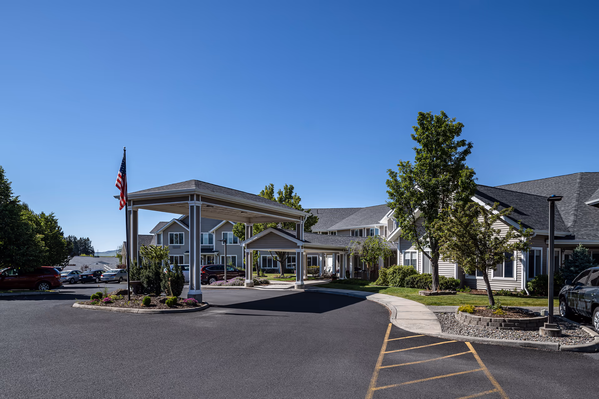 Exterior view of Bishop Place Senior Living facility on a clear sunny day, showing a covered entrance with an American flag, surrounding trees, parked cars, and a well-maintained driveway and landscaping.
