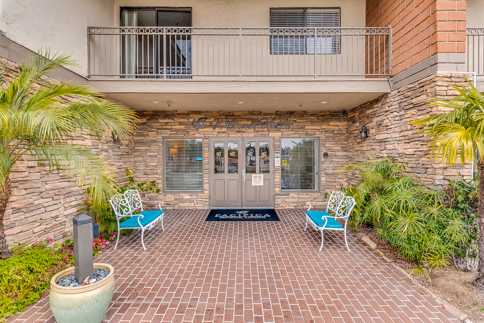Entrance to a senior living building with double glass doors set in a stone facade, two white benches with teal cushions, potted plants and a brick walkway.