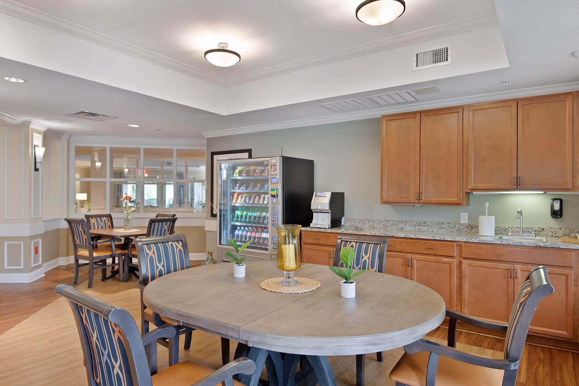A well-lit common area with a round wooden table surrounded by six chairs with patterned upholstery. On the table are two small potted plants and a decorative candle holder. In the background, there is a vending machine and a coffee machine on a countertop with wooden cabinets above and below. The room has hardwood flooring and soft, neutral-colored walls.