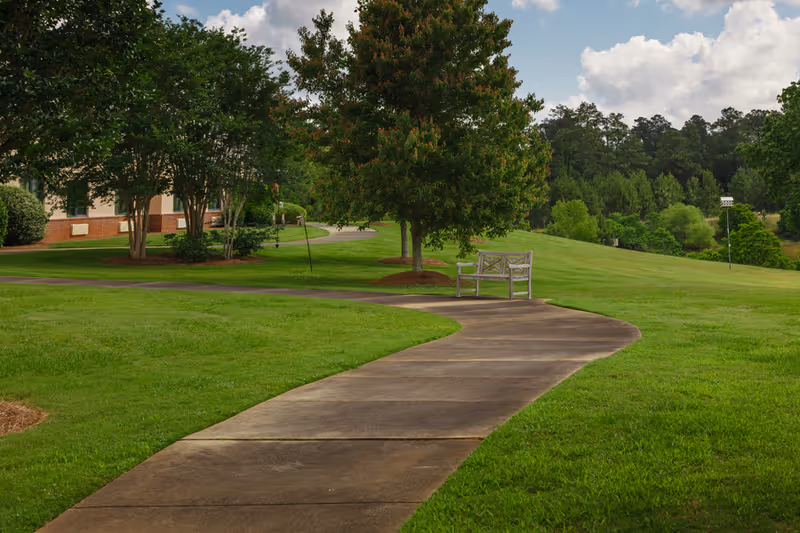 Curved concrete pathway through a well-maintained green lawn with trees and a wooden bench along the path. A building is partially visible on the left side, and the sky is partly cloudy.