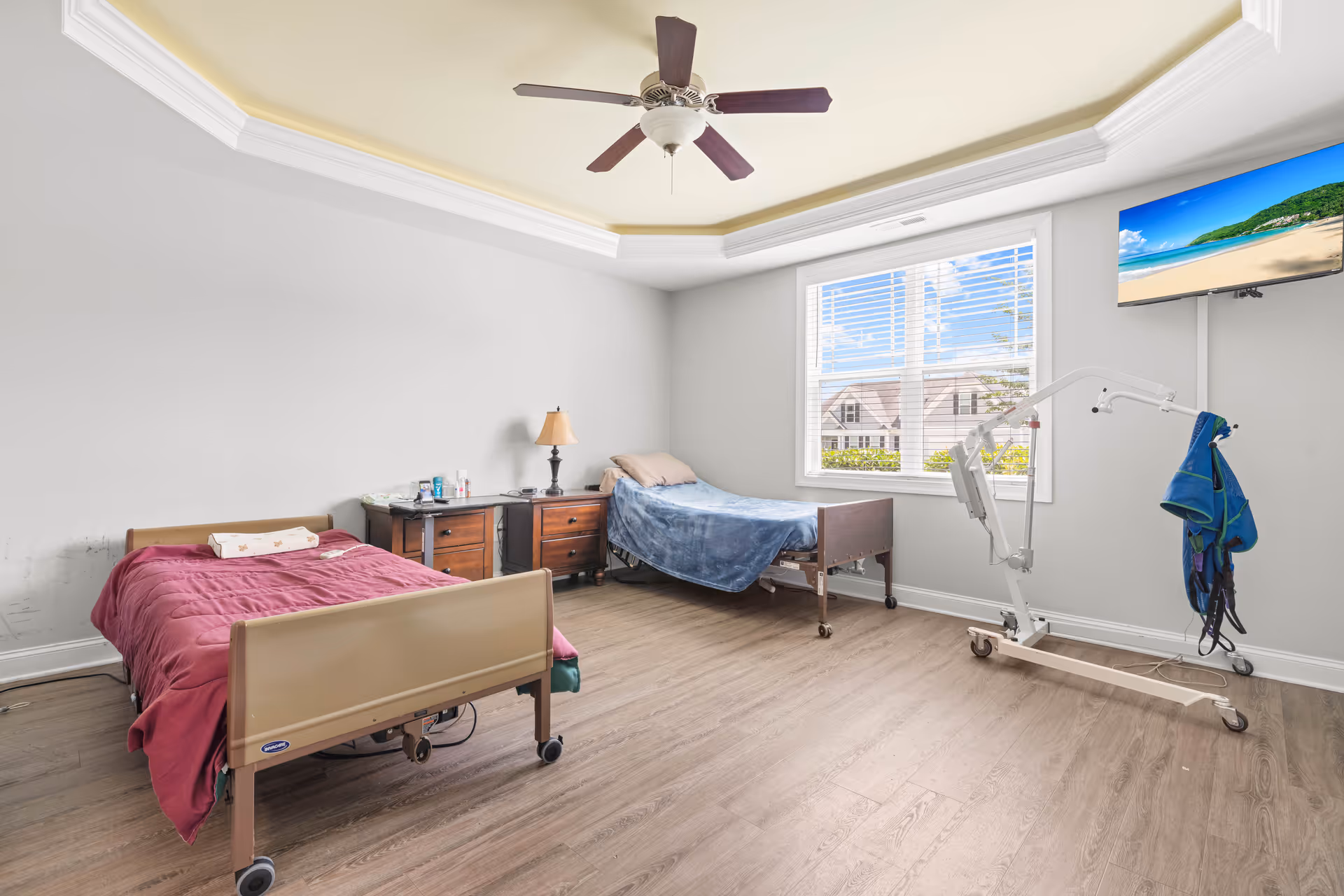Bright assisted-living bedroom with two hospital beds, bedside tables, ceiling fan, window, wall-mounted TV, and a patient lift.