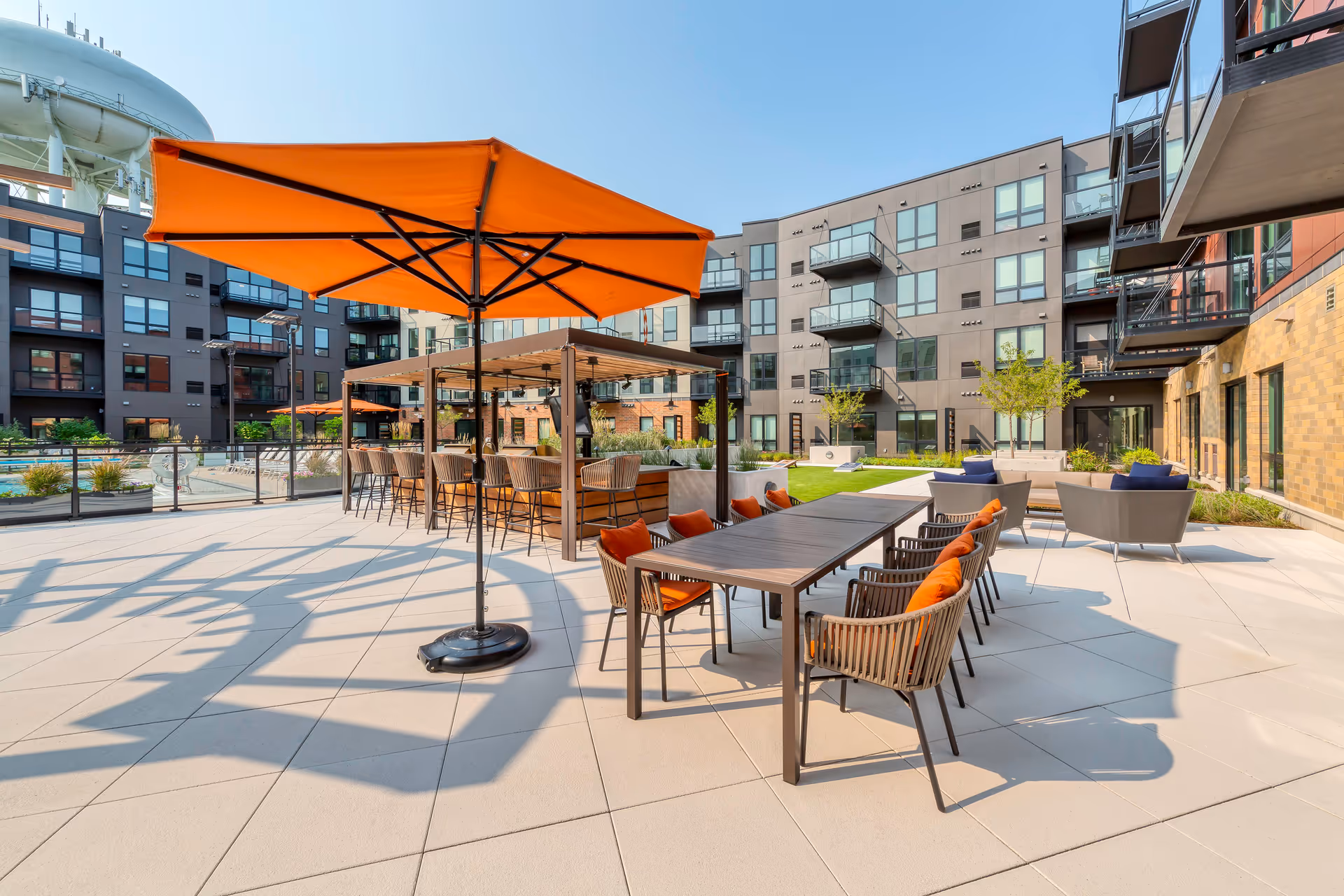 Outdoor patio area at Avidor Glenview featuring a large orange umbrella, a long table with chairs that have orange cushions, a pergola with bar stools, and modern apartment buildings in the background under a clear blue sky.
