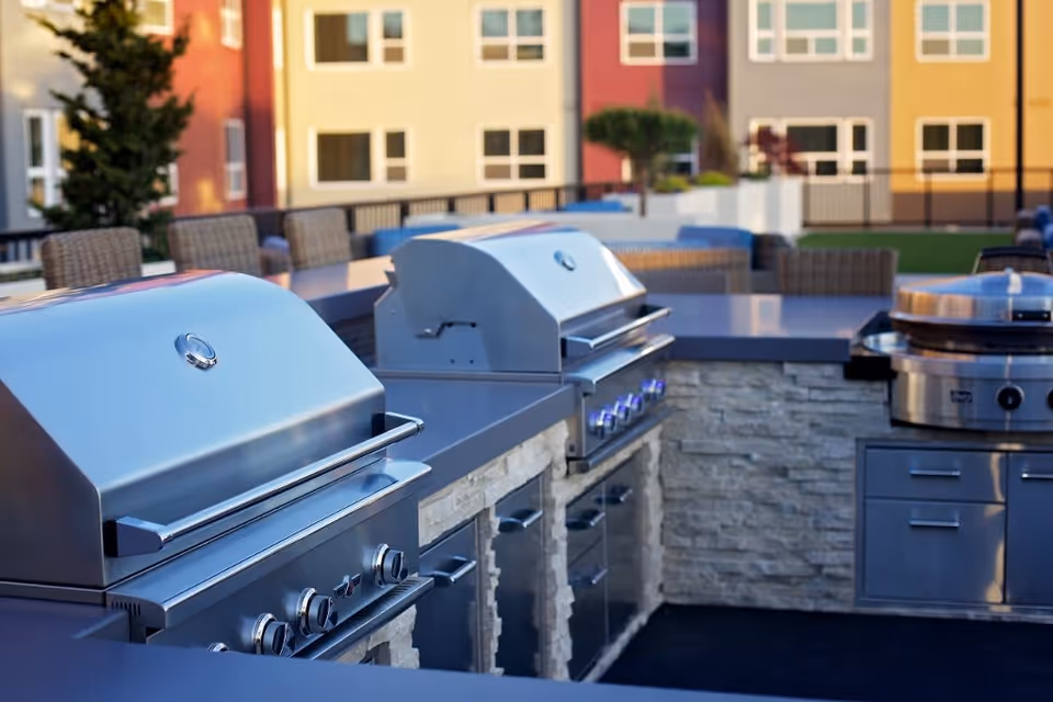 Outdoor grilling area with multiple stainless steel gas grills and a stone countertop, surrounded by wicker chairs and tables, with a colorful building facade in the background.