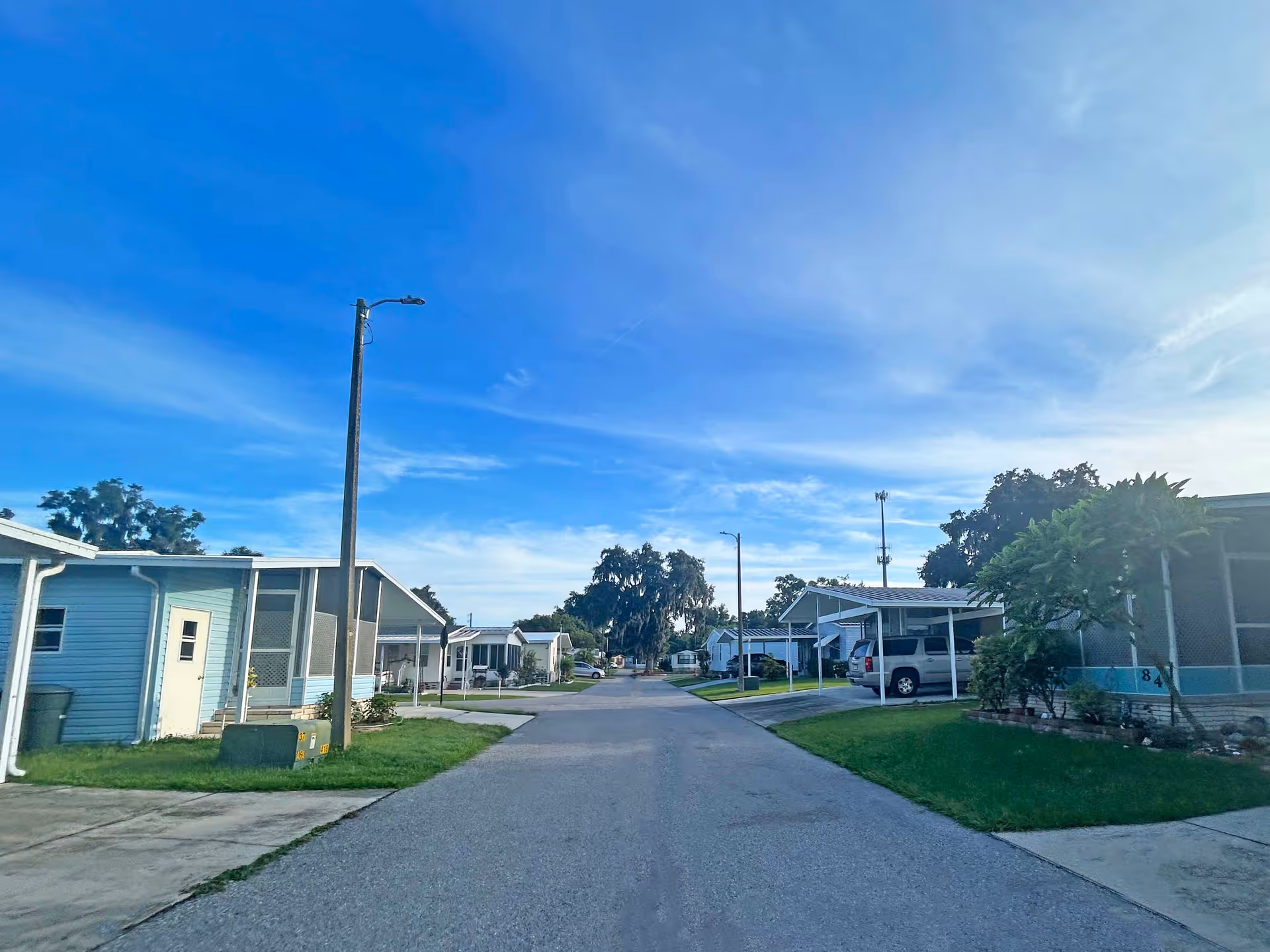 A wide residential street lined with small manufactured homes, lawns, driveways and parked cars under a bright blue sky.