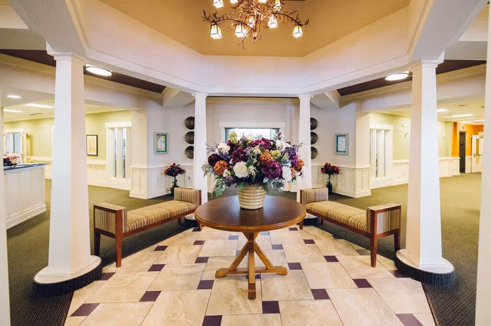 A bright and spacious interior common area with a round wooden table in the center holding a large floral arrangement. Two striped cushioned benches face each other on either side of the table. The space is framed by white columns and has a tiled floor with a geometric pattern. The surrounding area features carpeted floors, light-colored walls, and framed artwork.