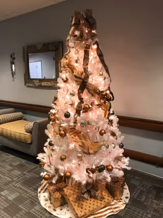 A decorated white Christmas tree with gold and bronze ornaments and ribbons, standing on a circular tree skirt with wrapped presents underneath. The tree is placed in a hallway with a cushioned bench and a mirror on the wall behind it.