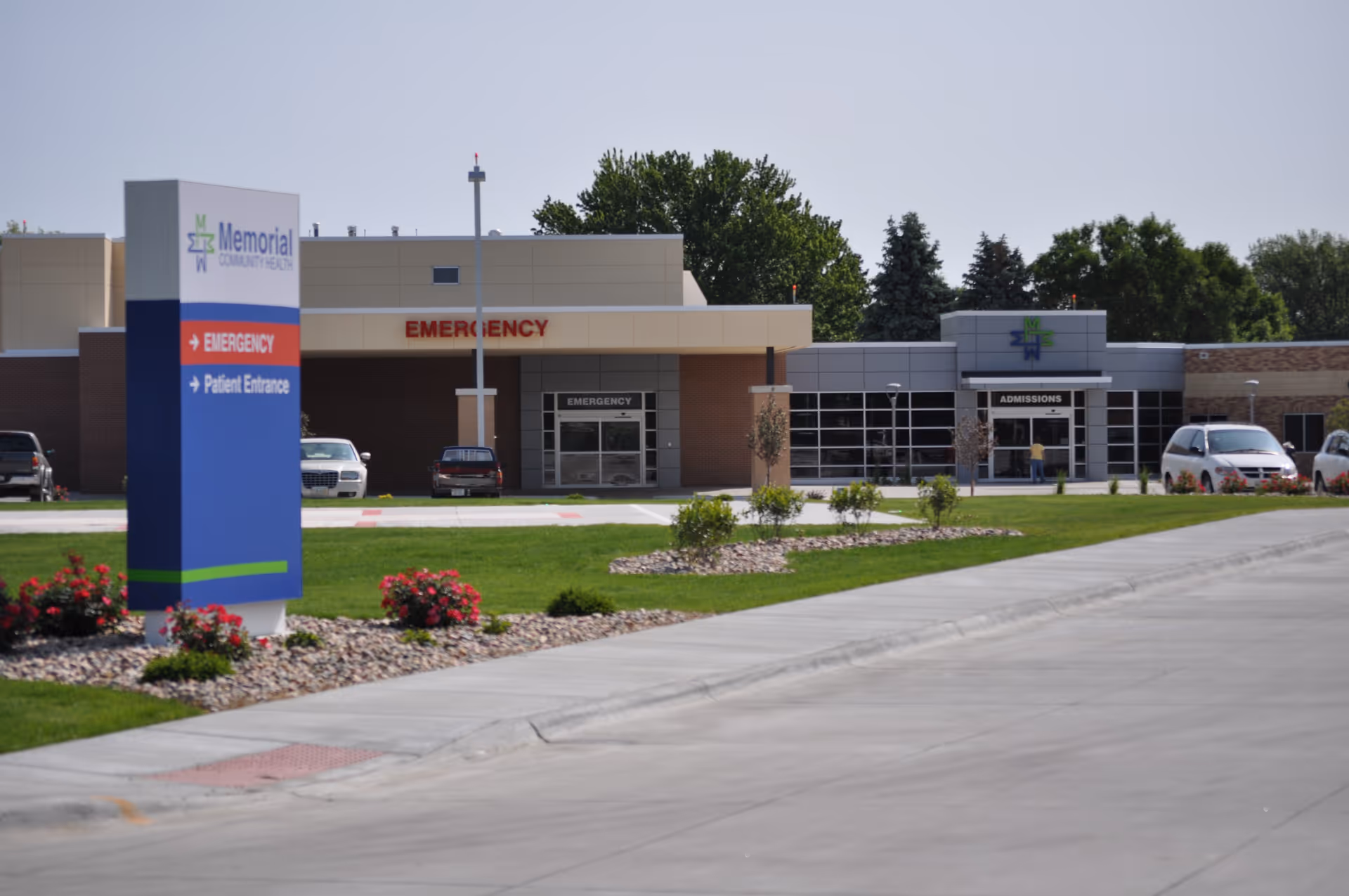 Exterior view of Memorial Community Health building showing the emergency entrance and admissions area with a sign in the foreground directing to Emergency and Patient Entrance. There are cars parked near the building and landscaping with grass, flowers, and small bushes.