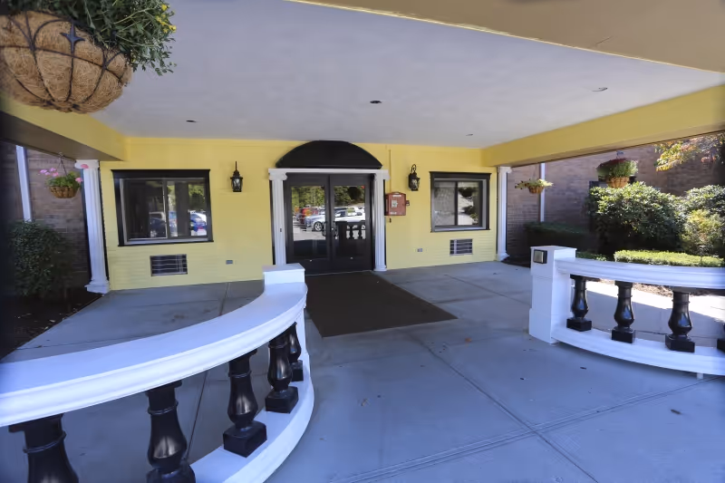 Covered entrance area of a building with yellow walls, black double doors, two windows, white decorative railings, hanging plants, and outdoor lighting fixtures.