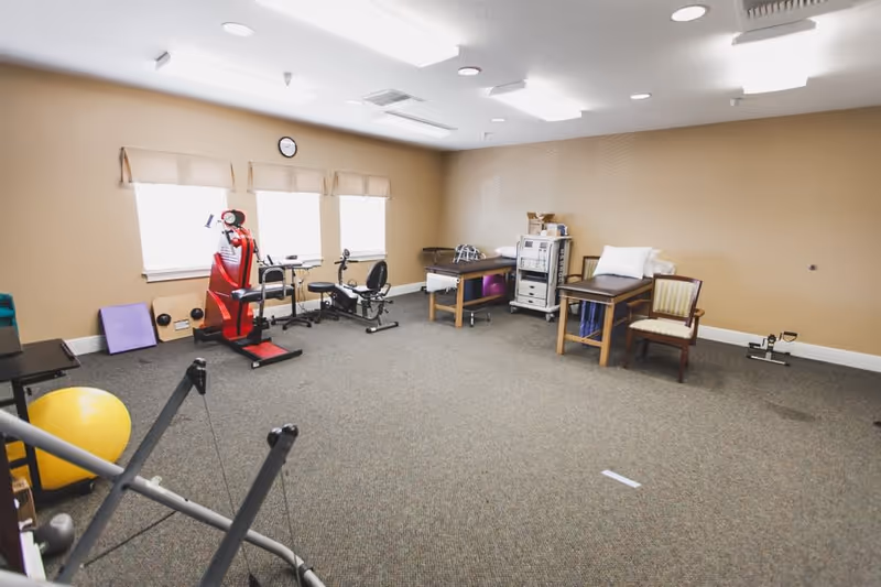 Bright interior therapy/exercise room with rehabilitation equipment, treatment tables, and chairs.