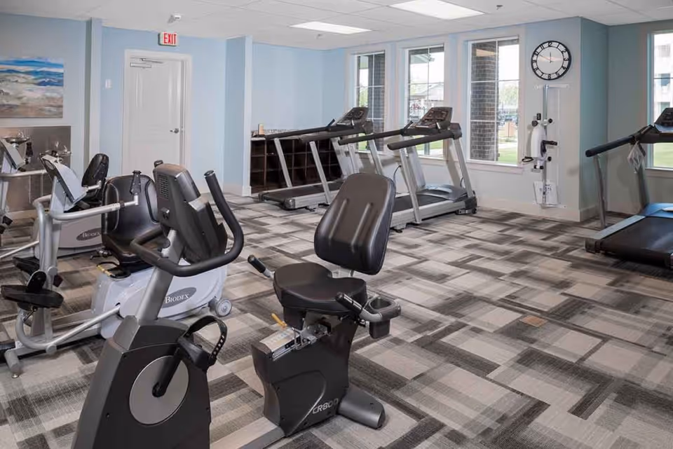A fitness room in a senior living facility with exercise equipment including stationary bikes, treadmills, and a wall-mounted exercise machine. The room has large windows allowing natural light, light blue walls, a patterned carpet, and a clock on the wall.