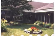 Grassy courtyard featuring a circular stone-bordered flower bed, trees and shrubs in front of a single-story building with large windows and a covered walkway.