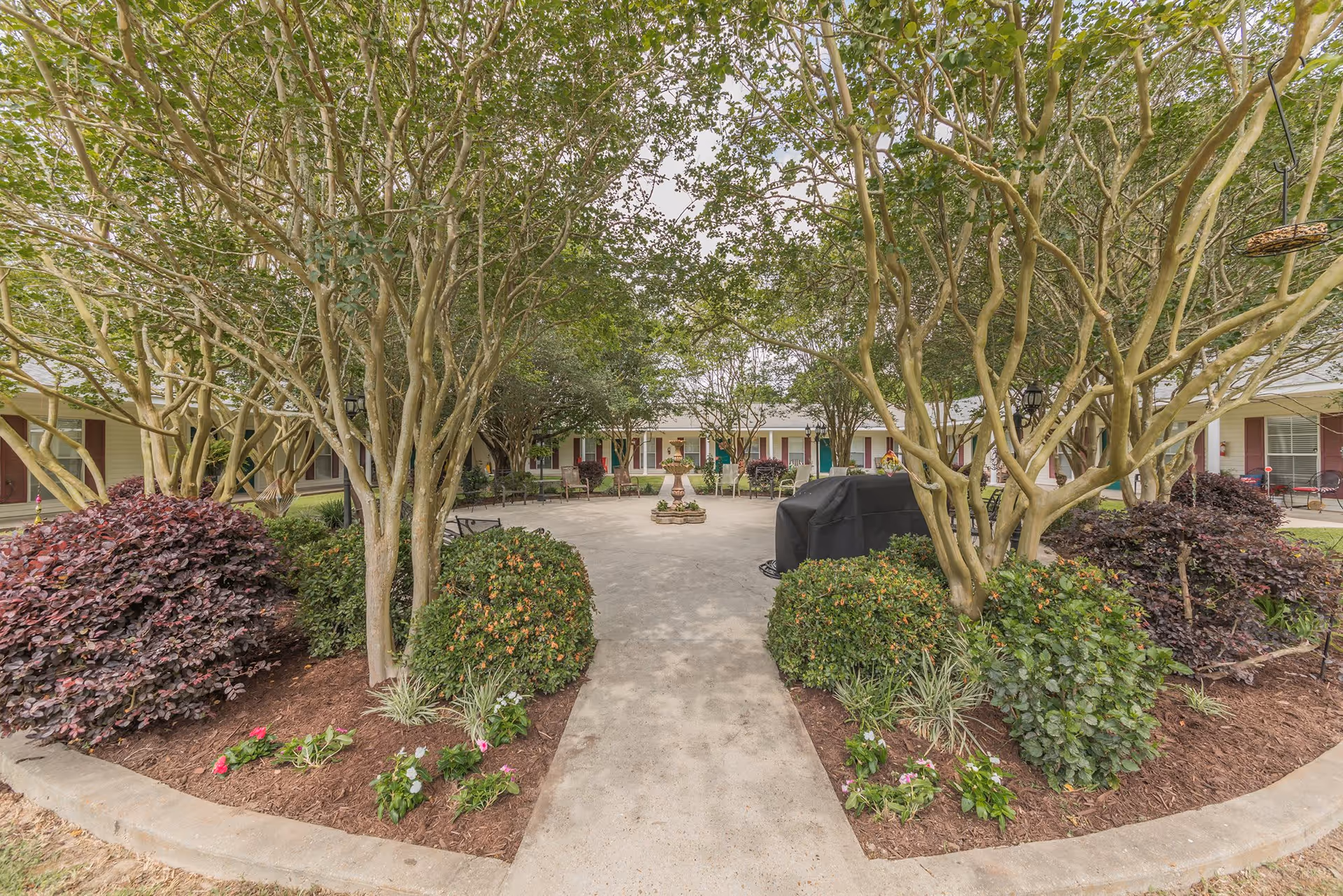 A peaceful outdoor courtyard area at Rosewood Assisted Living featuring a concrete pathway lined with well-maintained bushes and flowering plants. Several trees provide shade, and there are benches and chairs arranged around the courtyard. A covered grill is visible on the right side, and the building with multiple doors and windows surrounds the courtyard.