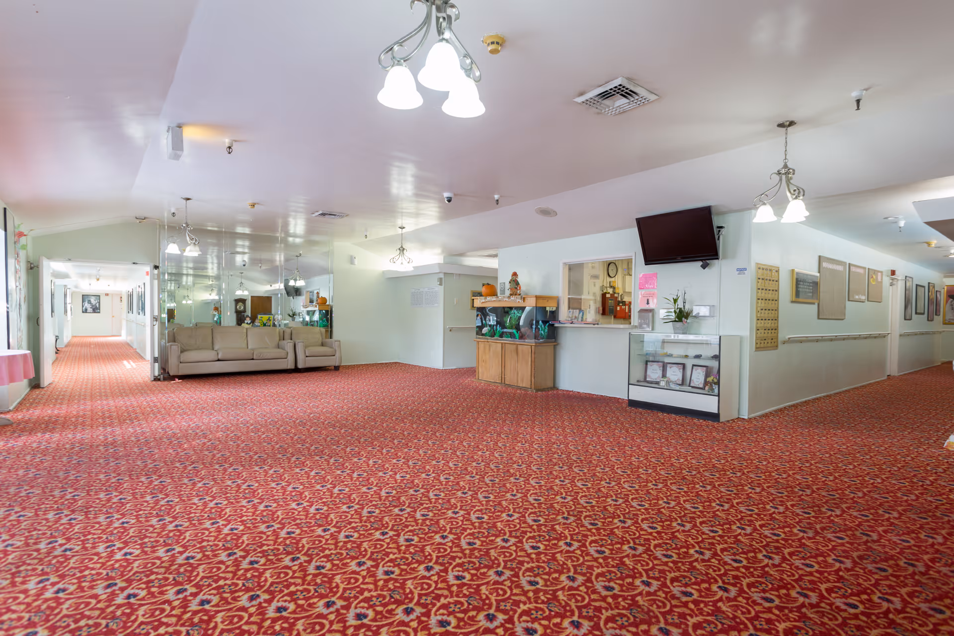 A spacious hallway in a senior living facility with red patterned carpet, beige couches along the left wall, a fish tank on a wooden stand, a wall-mounted TV, and various framed pictures and bulletin boards on the walls. The ceiling has multiple light fixtures and ventilation panels.