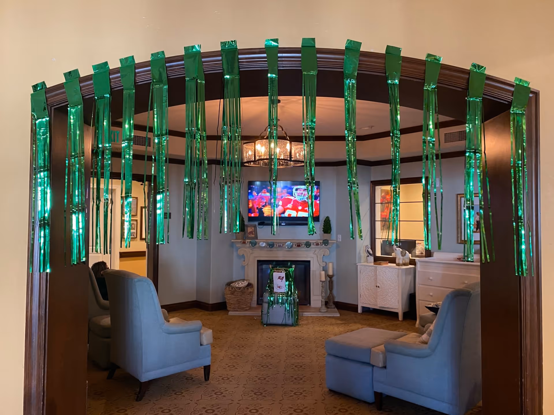 A cozy living room decorated with green hanging streamers. The room features light blue armchairs, a fireplace with a TV mounted above it showing a football game, and a white cabinet with decorative items. The walls are light-colored with dark wood trim.