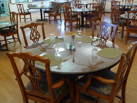 A dining room with multiple wooden tables and chairs. The tables are set with placemats, upside-down drinking glasses, and silverware. The floor is wooden, and the room is well-lit with natural light.