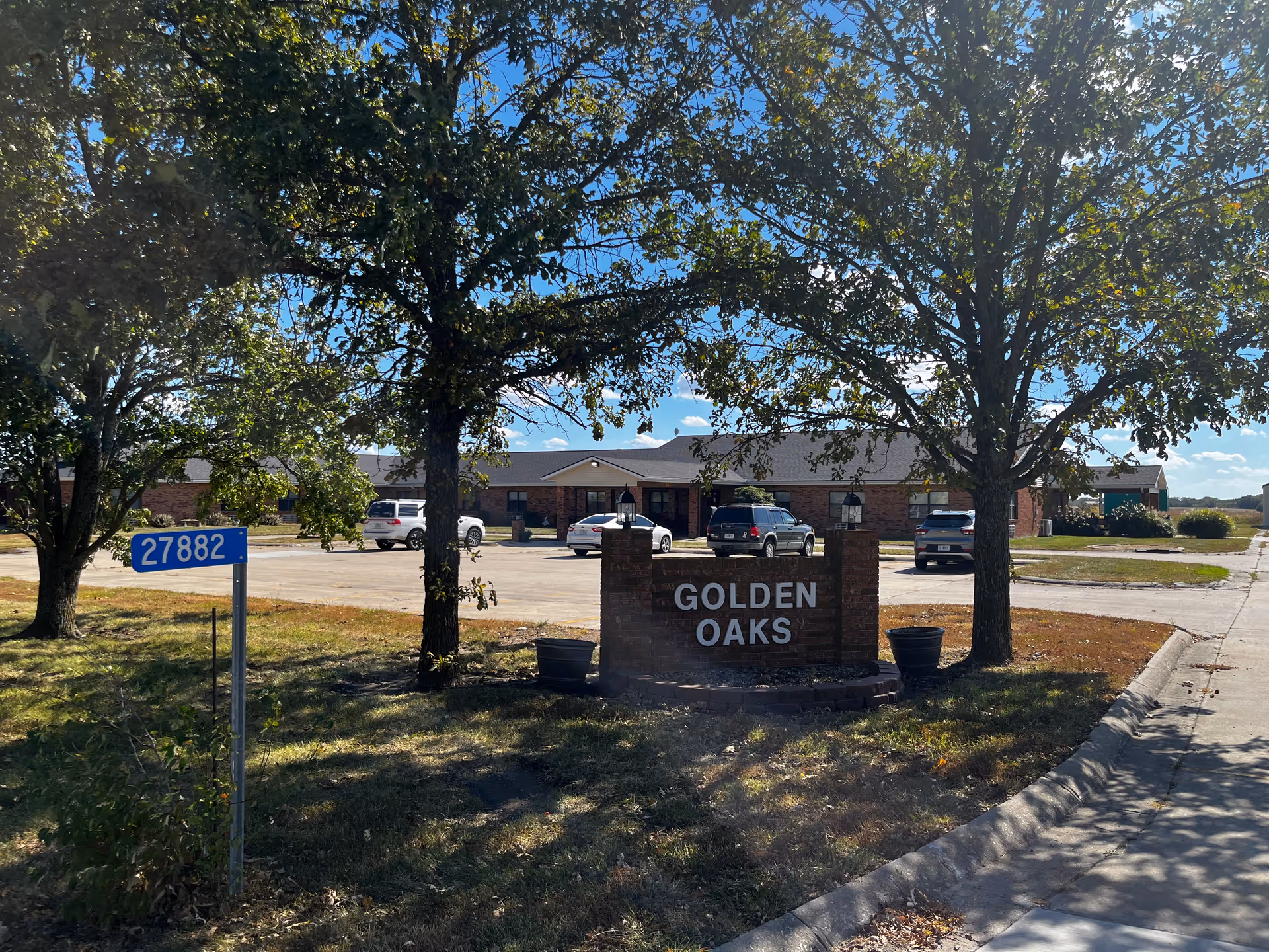 Exterior view of Golden Oaks Assisted Living facility with a brick sign displaying the name 'GOLDEN OAKS' in front. The building is a single-story structure with a parking lot containing several cars. Trees with green leaves partially shade the sign and the area around it. A blue street number sign with the number 27882 is visible on the left side of the image.