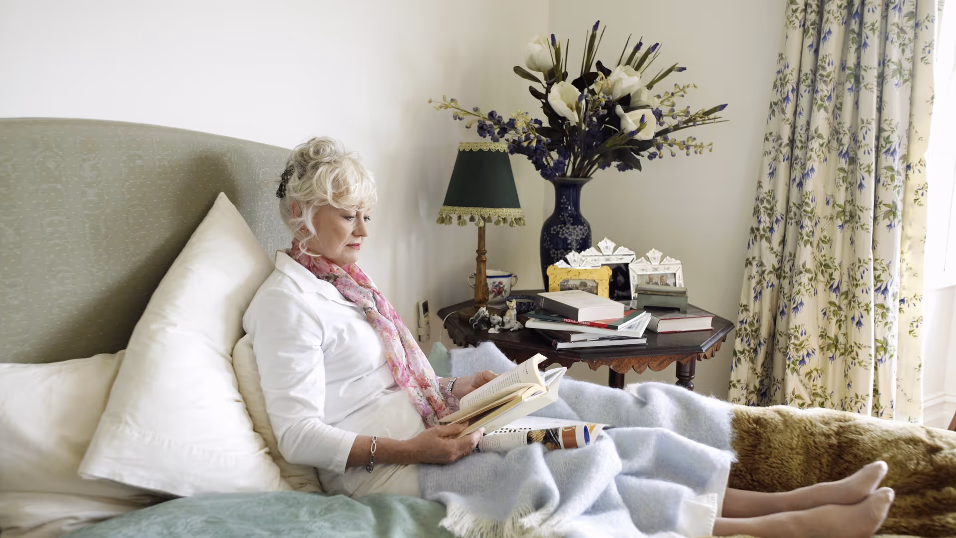 An older woman sits propped up in bed reading a book beside a table with a lamp, books, and a vase of flowers, with floral curtains in the background.