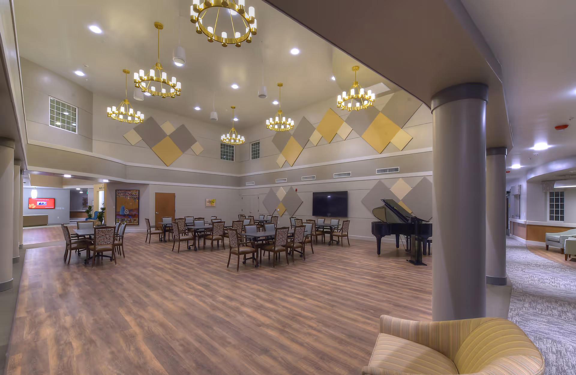 Spacious senior living common room with dining tables and chairs, chandeliers, a grand piano, and decorative wall panels.