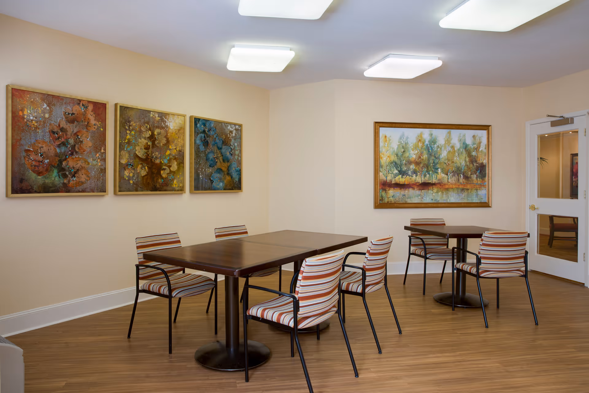 A well-lit room with wooden flooring featuring two dark wood tables surrounded by striped cushioned chairs. The walls are decorated with four framed paintings, three with floral designs and one depicting a landscape with trees and a body of water. A white door with a glass panel is visible on the right side of the room.