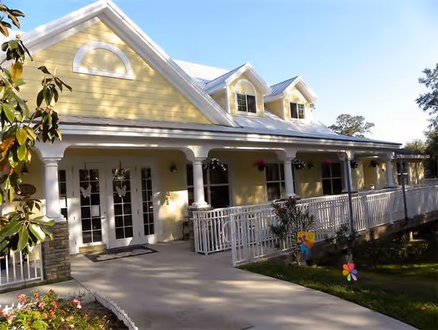 Front exterior view of a yellow assisted living facility building with white trim, a metal roof, and a covered porch with white railings and columns. There is a concrete walkway leading to double glass doors, surrounded by greenery and flowers.
