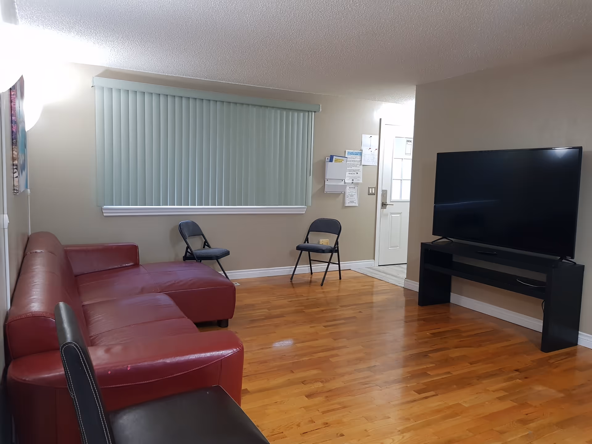 A living room with a red leather sectional sofa, two black folding chairs, a large flat-screen TV on a black TV stand, hardwood floors, beige walls, and a window with vertical blinds.