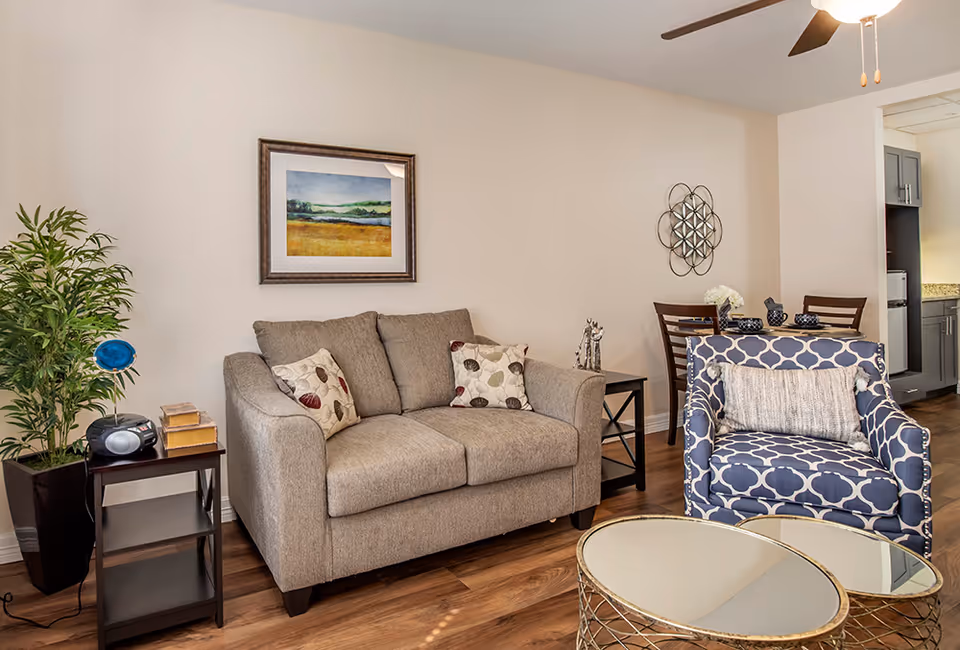 A cozy living room area with a beige loveseat adorned with two patterned pillows, a blue and white patterned armchair with a gray cushion, and two round glass-top coffee tables with gold metal bases. To the left is a small dark wooden side table with a radio and some books, next to a tall green potted plant. On the wall above the loveseat is a framed landscape painting. In the background, there is a small dining area with a wooden table and chairs, and a glimpse of a kitchen with gray cabinets and granite countertops.