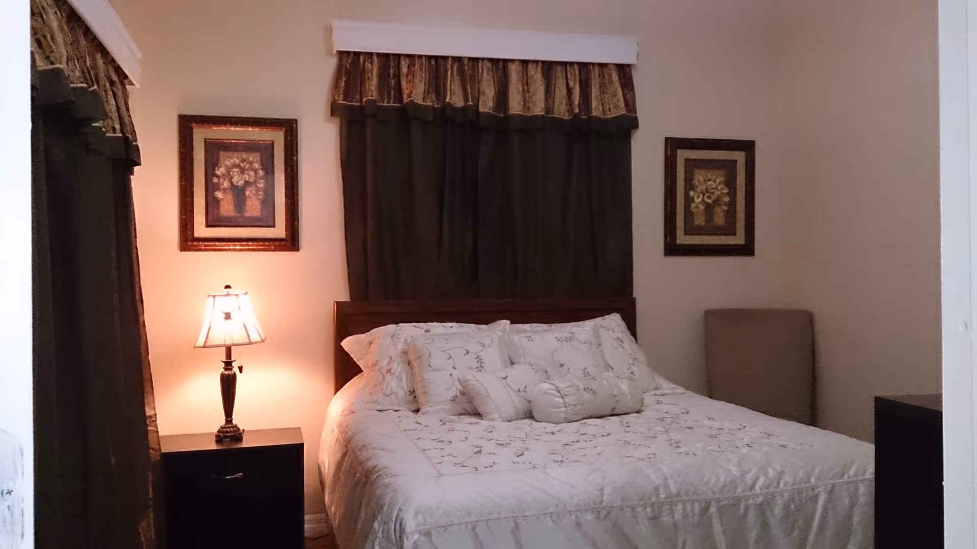 A cozy bedroom with a bed covered in white floral bedding and multiple pillows. There are dark curtains with a valance above the bed, two framed floral pictures on the wall, a bedside table with a lamp emitting warm light, and a cushioned chair in the corner.