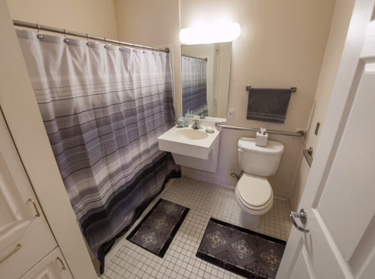 Compact bathroom with a striped shower curtain, wall-mounted sink and mirror, toilet with grab bars, and tiled floor with two mats.
