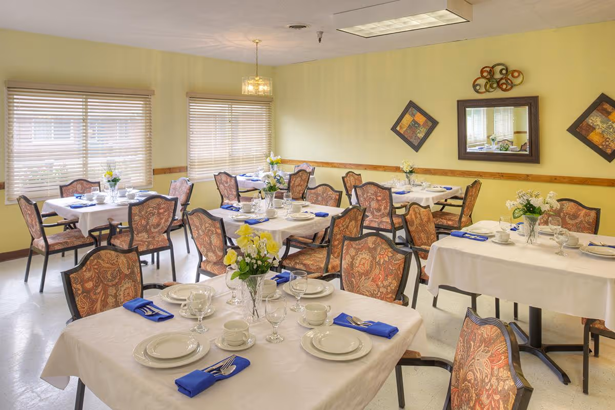 A dining room with several tables covered in white tablecloths, each set with plates, cups, glasses, and blue napkins. The chairs have patterned upholstery. There are windows with blinds, wall decorations including a mirror and framed art, and floral centerpieces on the tables.