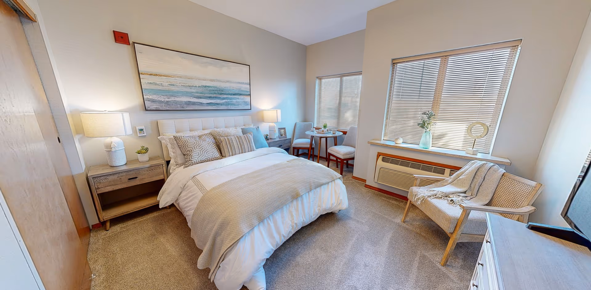 A bright, neatly staged bedroom with a made bed, nightstands, a small table and chairs by the windows, and a cozy armchair.