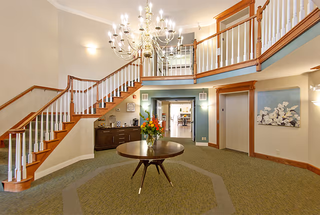Spacious interior lobby area with a large wooden staircase featuring white balusters, a round wooden table with a vase of flowers in the center, a chandelier hanging from the ceiling, and a hallway leading to another room.