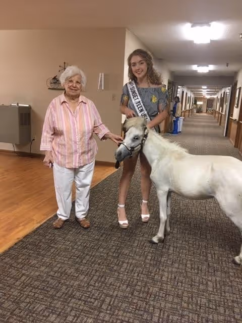 An elderly woman and a young woman wearing a sash stand in a hallway of a retirement community. The young woman is holding a small white pony on a leash. The hallway has carpeted flooring and beige walls with framed pictures and lighting fixtures on the ceiling.