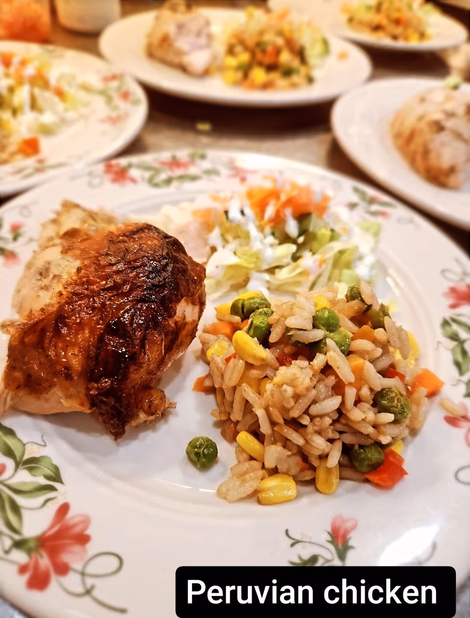 A plate of Peruvian chicken served with a side of mixed vegetable rice and a small salad with lettuce and shredded carrots. The plate has a floral pattern around the edge. In the background, there are other plates with similar food items.