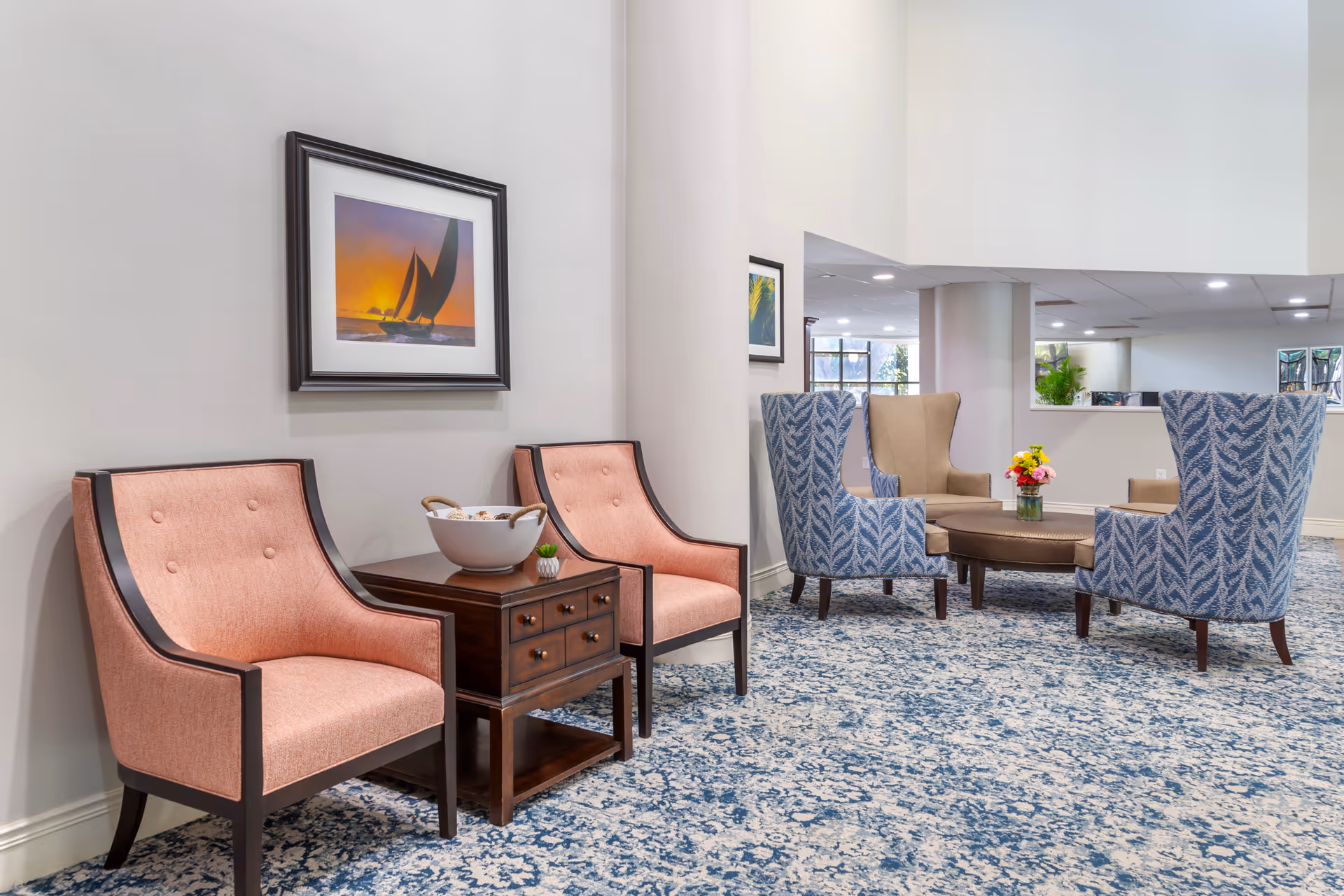 A seating area in a senior living facility with two pink upholstered chairs flanking a small wooden table with decorative items, and a group of four blue patterned and beige armchairs arranged around a round coffee table with a flower vase. The room has a patterned blue and white carpet, light-colored walls, and framed artwork on the walls.