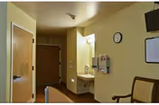 Interior view of a room in a care center showing a wooden door, a small sink with a mirror above it, a clock on the wall, a mounted television, a whiteboard, and a chair with wooden armrests.