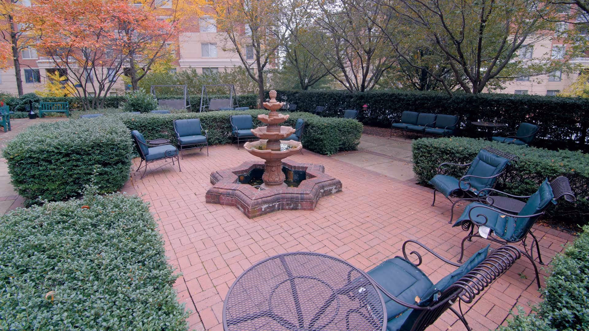 Outdoor courtyard area with a central multi-tiered brick fountain surrounded by green cushioned metal chairs and benches. The space is paved with red bricks and bordered by neatly trimmed hedges and trees with autumn foliage. Apartment building windows are visible in the background.