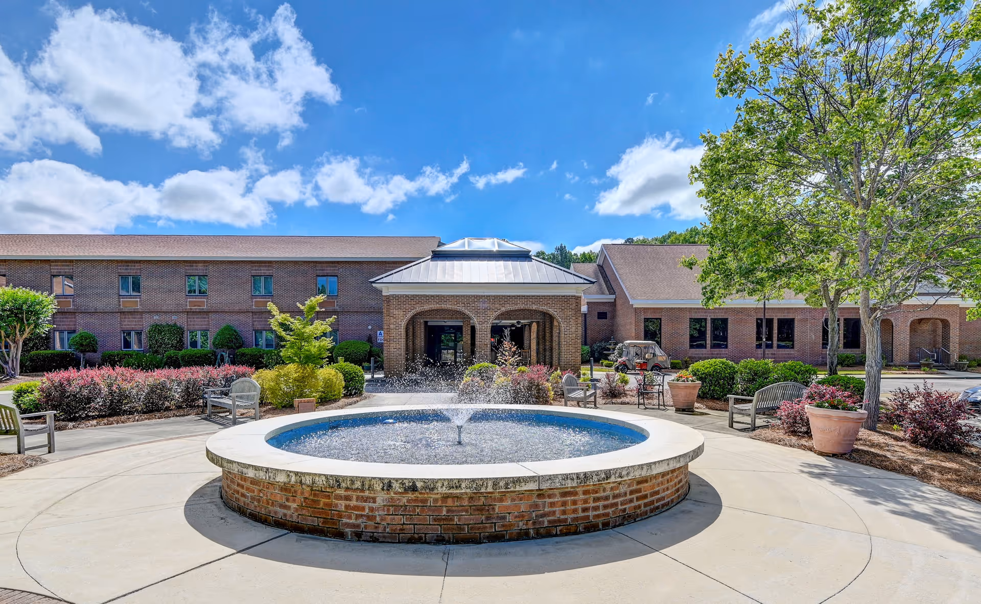 Brick senior living building front with a circular fountain and landscaped courtyard under a blue sky.