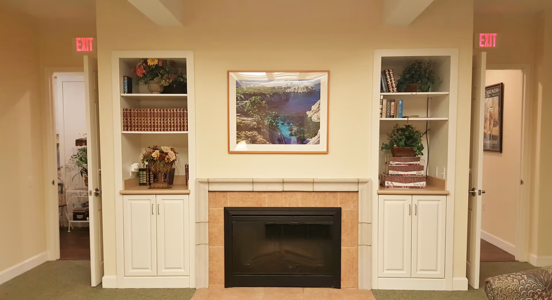Interior view of a cozy room with a fireplace in the center, flanked by two built-in white shelves filled with books, plants, and decorative items. Above the fireplace hangs a framed picture of a coastal landscape. Two open doorways with exit signs above them are visible on either side of the shelves.