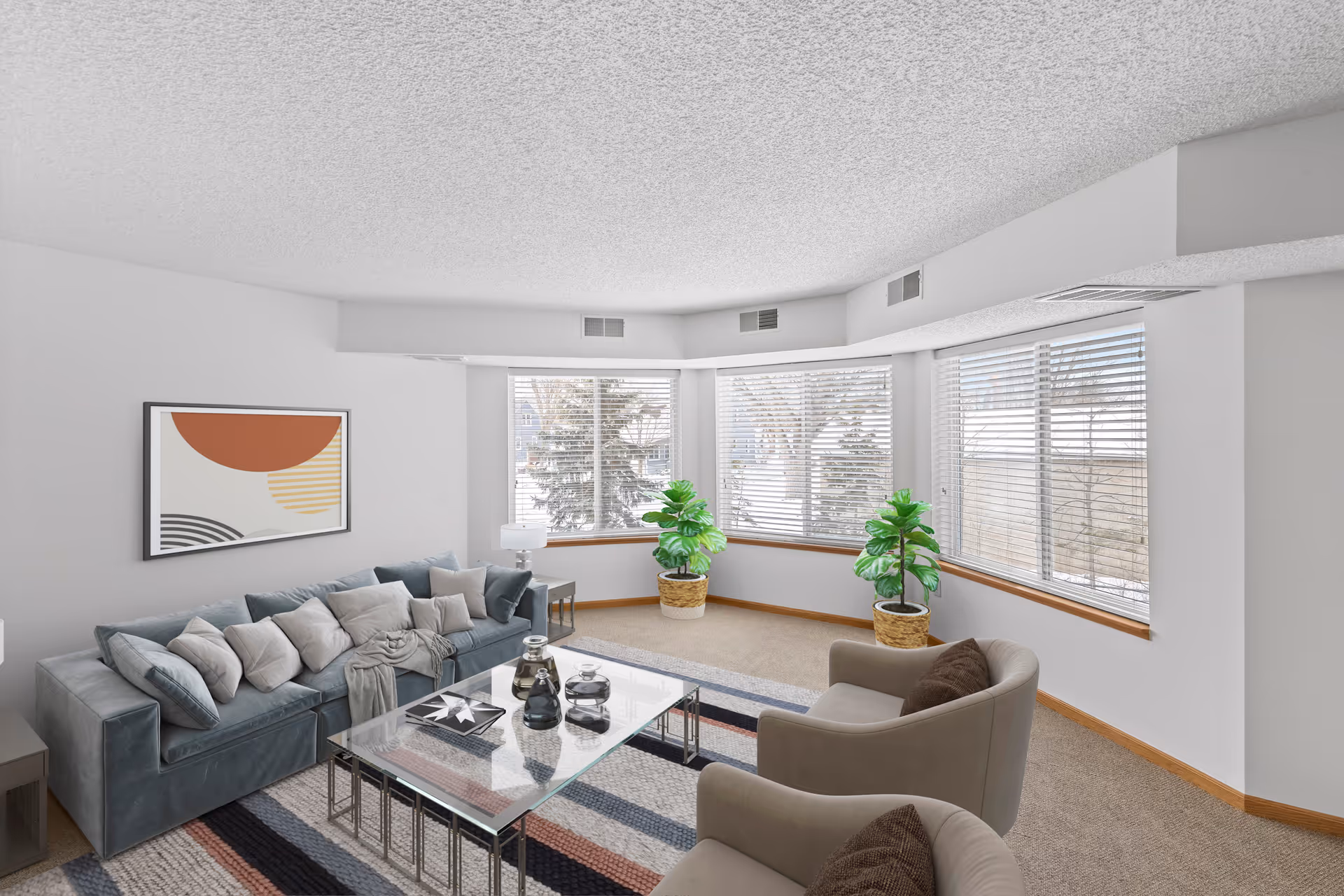 A bright living room with a large blue sofa adorned with multiple cushions, two beige armchairs with brown pillows, a glass coffee table with decorative items, a modern abstract painting on the wall, and three large windows with white blinds letting in natural light. Two potted plants are placed near the windows, and the room has light-colored walls and carpeted flooring.