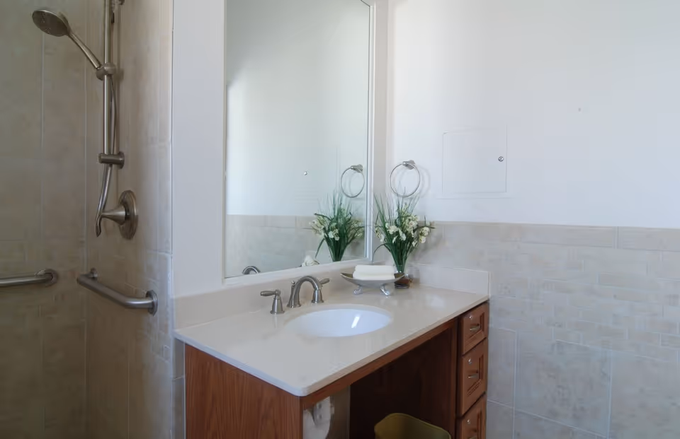 A clean bathroom with a beige tiled shower area featuring a handheld showerhead and grab bars. A countertop with an under-mount sink, silver faucet, and a large mirror above it. On the countertop, there is a small vase with white flowers and a soap dish with a bar of soap. The walls are partially tiled with beige tiles and painted white above.