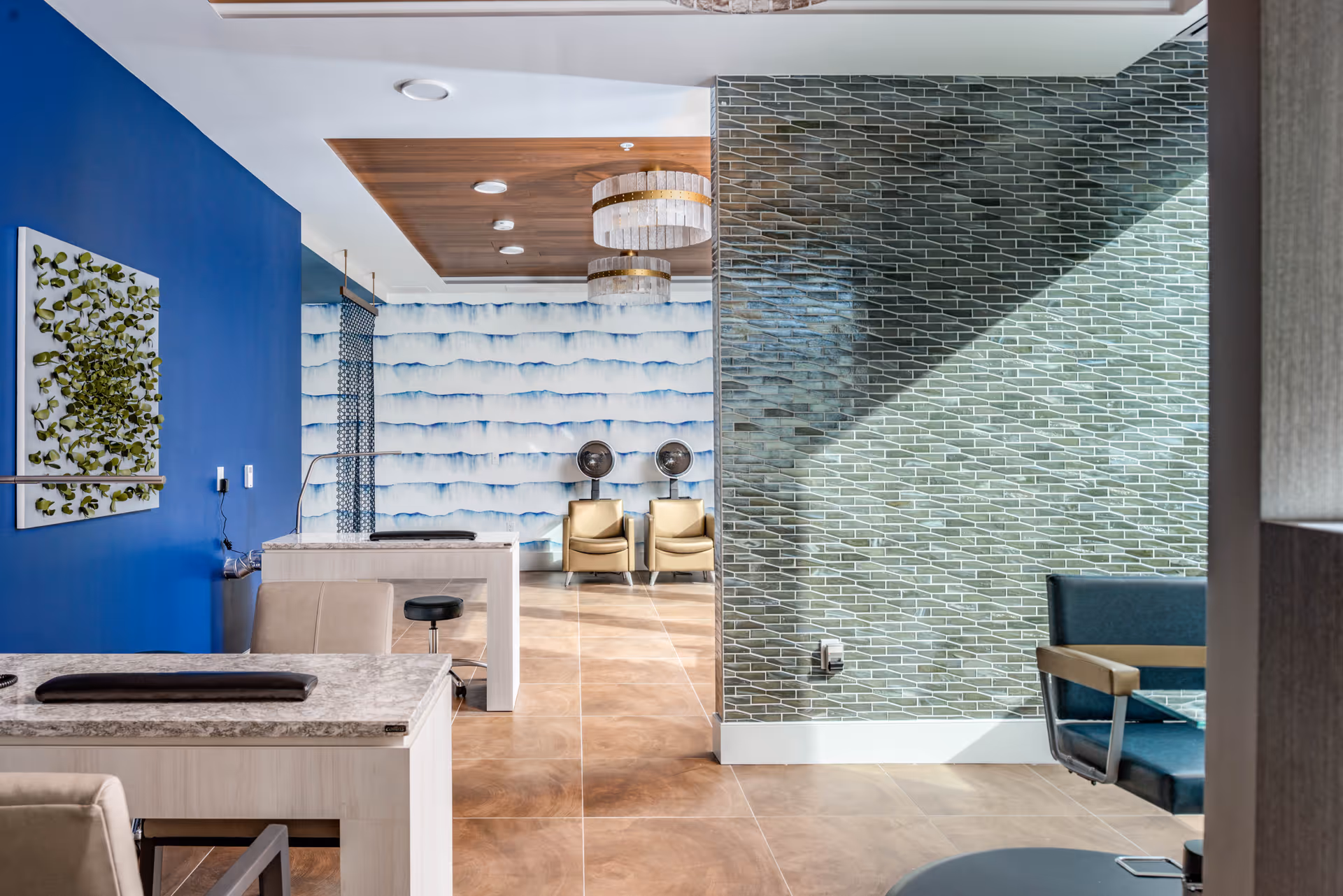 Interior view of a modern salon area with beige chairs, two hair dryers against a blue and white patterned wall, a blue accent wall with a green leaf artwork, marble countertops, and a green tiled wall with sunlight casting shadows.