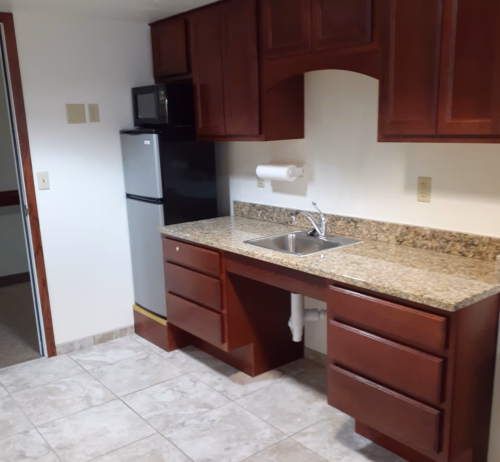 A small kitchen area with dark wood cabinets, a granite countertop with a stainless steel sink, a paper towel holder mounted on the wall, a compact refrigerator, and a microwave on top of the refrigerator. The floor is tiled with light-colored tiles.