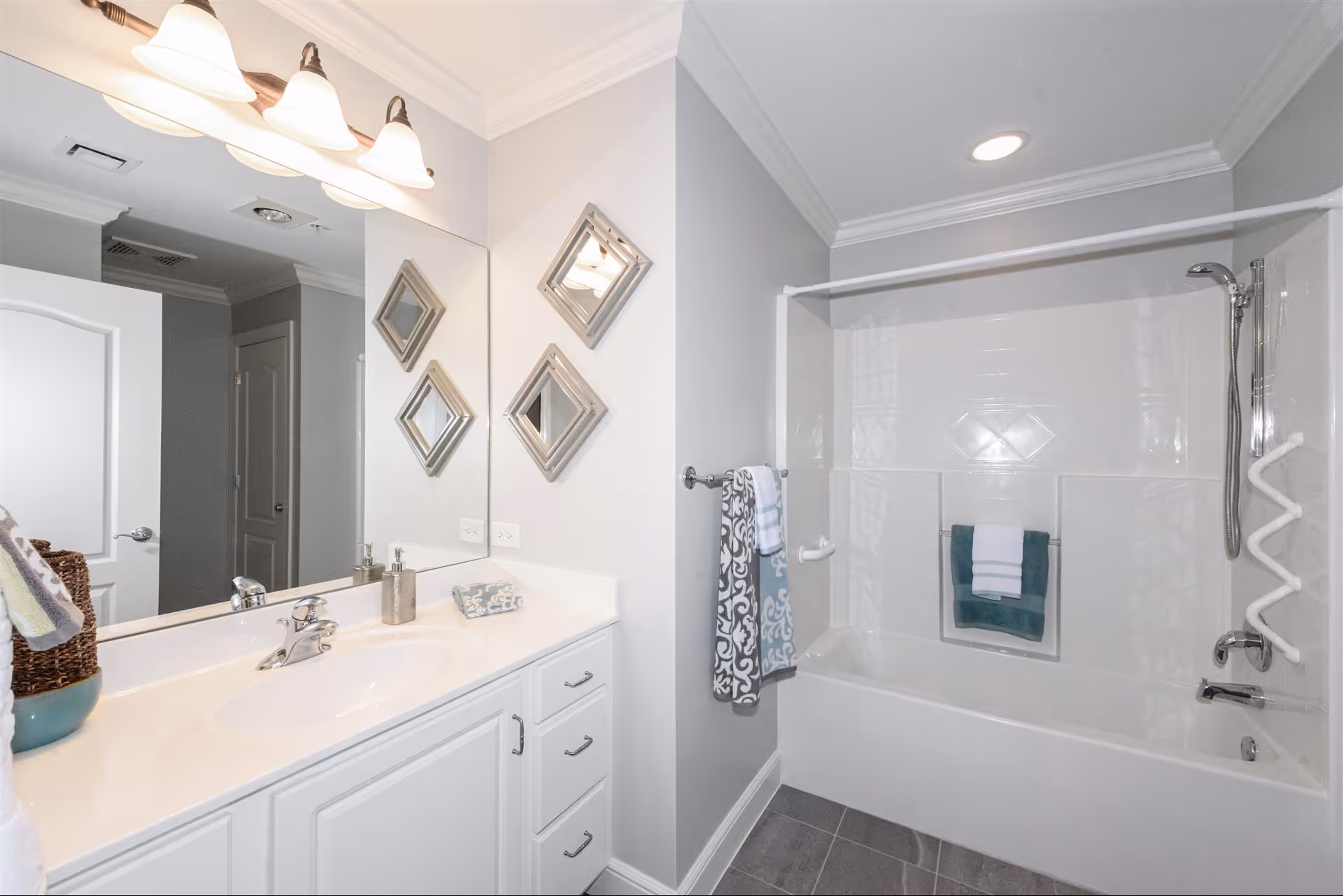 A clean and modern bathroom featuring a white vanity with a sink, a large mirror above it, and three decorative square mirrors on the adjacent wall. The bathroom also includes a white bathtub with a showerhead, a towel rack holding two towels, and a white grab bar for safety. The walls are painted light gray, and the floor has gray tiles.