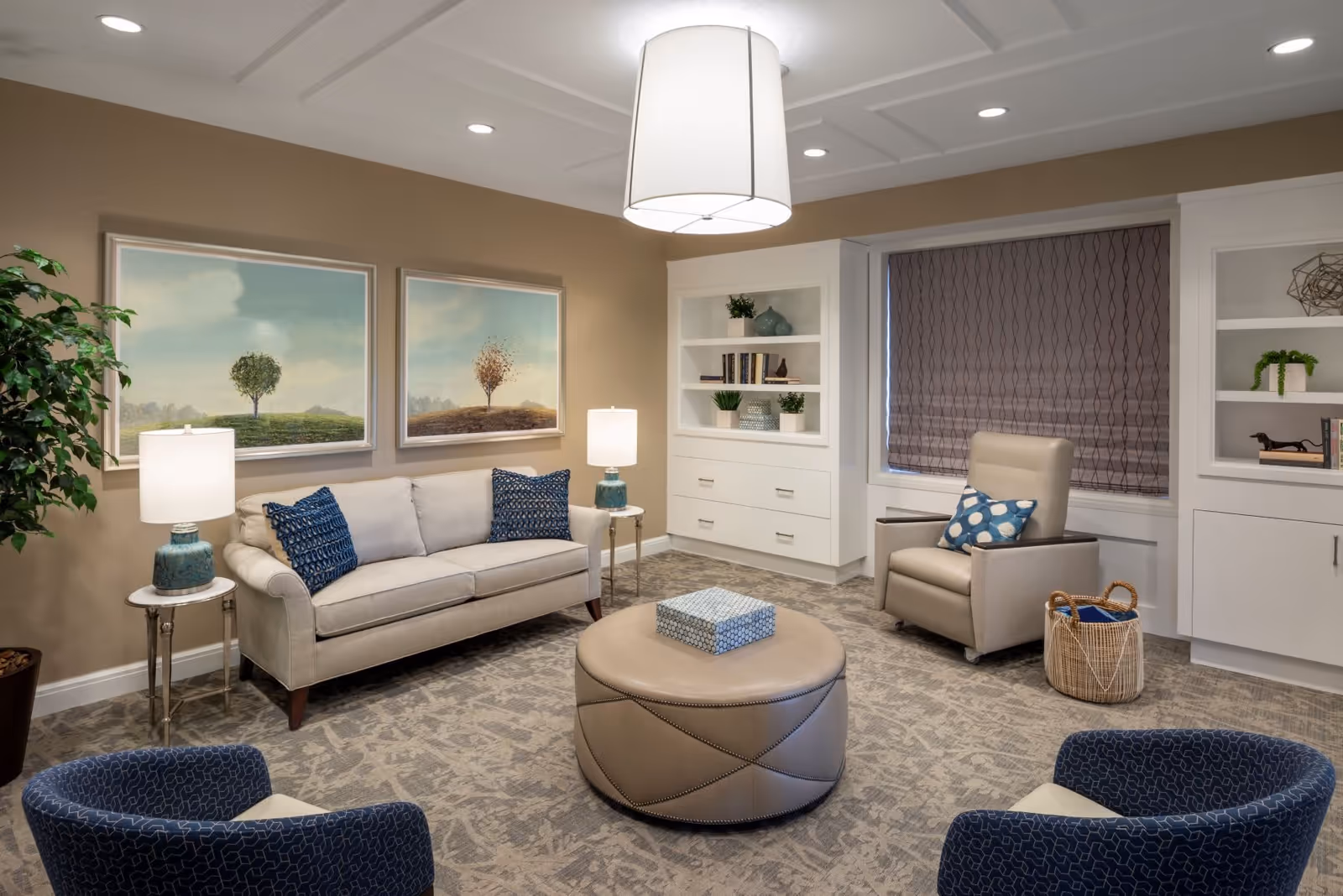 A cozy living room with a beige sofa adorned with blue patterned pillows, two blue armchairs, a beige leather ottoman with a decorative box on top, a beige armchair with a blue and white pillow, and built-in white shelves with plants and books. The walls are painted light brown and decorated with two framed paintings of trees on hills. A large white pendant light hangs from the ceiling, and there are two table lamps on either side of the sofa. A basket with a blue blanket is placed near the armchair.