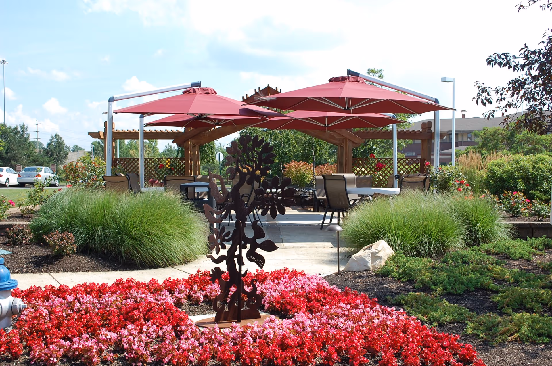 Outdoor garden area with vibrant red and pink flowers in the foreground, green shrubs, and a decorative metal sculpture. In the background, there are two large red umbrellas shading tables and chairs under a wooden pergola, with a clear blue sky above.