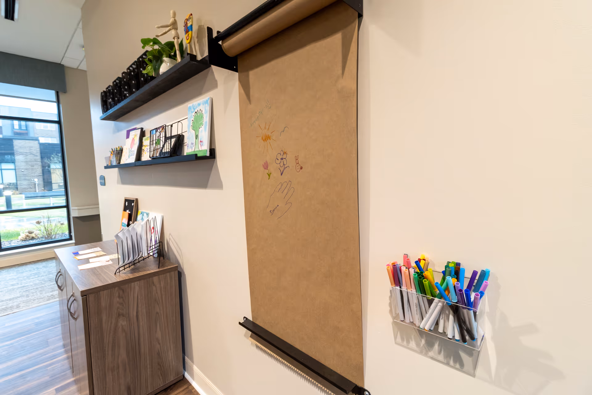 A creative corner in a room with a roll of brown craft paper mounted on the wall, partially drawn with colorful markers. Next to it is a clear container holding various colored markers. On the adjacent wall, two black shelves display art supplies, small plants, and children's drawings. Below the shelves is a wooden cabinet with papers and folders on top. A large window shows an outdoor view of a building and greenery.
