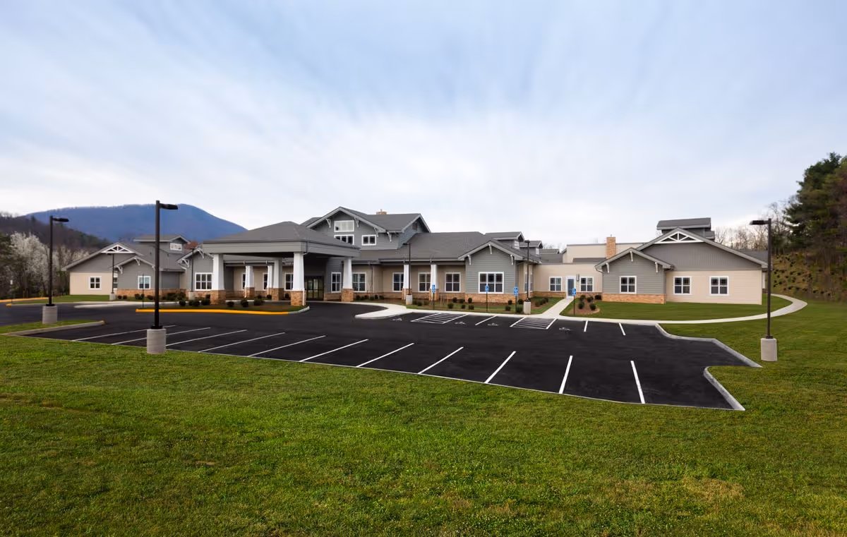 Exterior view of Friendship Assisted Living facility showing a large single-story building with a covered entrance, surrounded by a parking lot and green lawn, with mountains visible in the background under a cloudy sky.