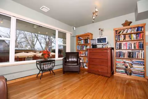 A cozy living room in a senior facility with bookshelves, leather chair, and wooden floor.